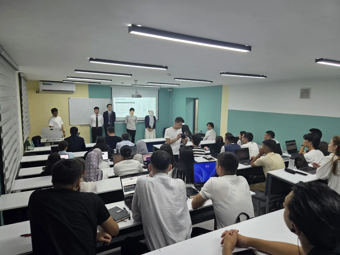 Young Joon Lee and the four tutors of the KOICA-TIU AI Training Center's third cohort standing at the front of Room 411 under a "Meet your team" slide, with students seated at their laptops, during the September 2026 orientation at Tashkent International University