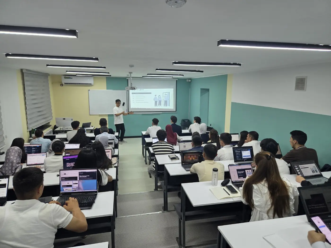 A wide view of Room 411 from the back of the room as students follow the "Program at a glance" slide during the Cohort 3 orientation