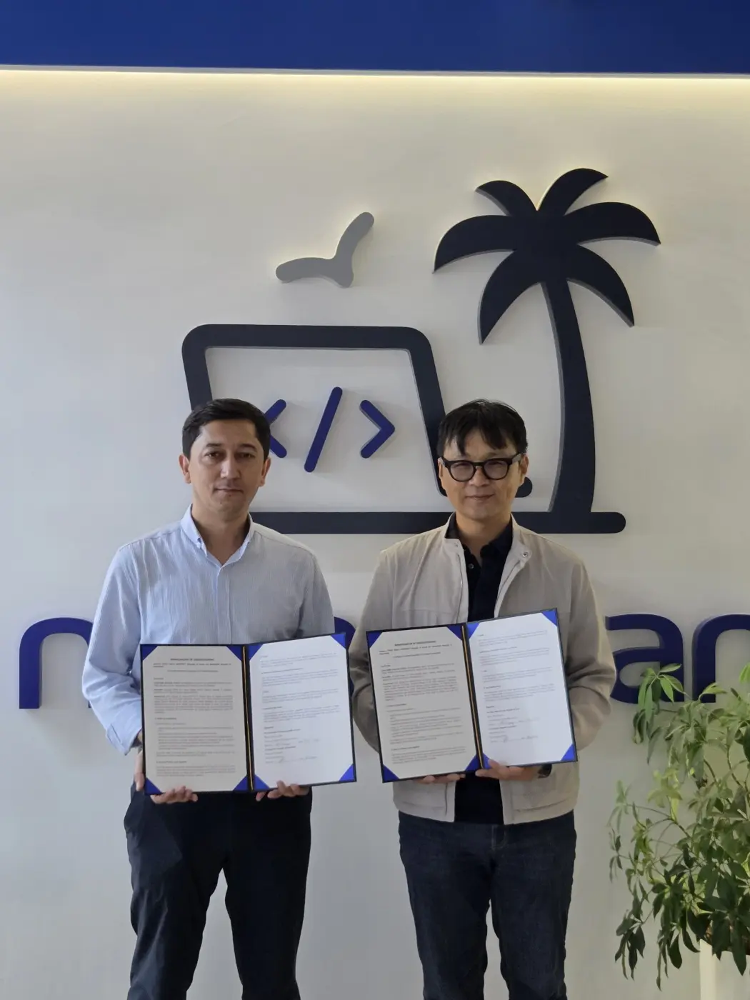 Young Joon Lee of Cheju Halla University and Mamur Djurayev, Managing Director of Nomadian, holding the signed MOU folders in front of the Nomadian logo wall in Tashkent